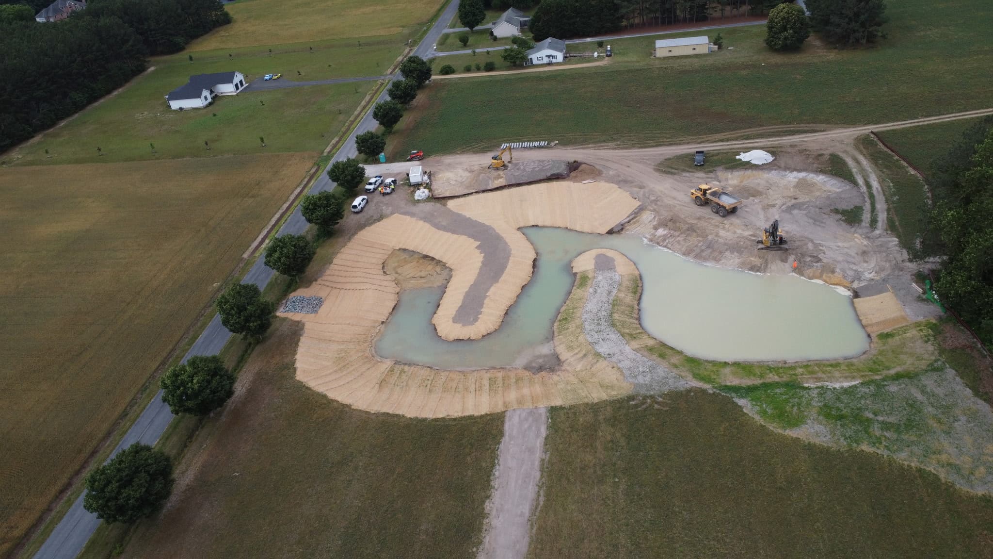 Aerial view of the engineered treatment wetland from the opposite end, showing the serpentine flow path and freshly stabilized banks alongside the roadway and adjacent farm fields.