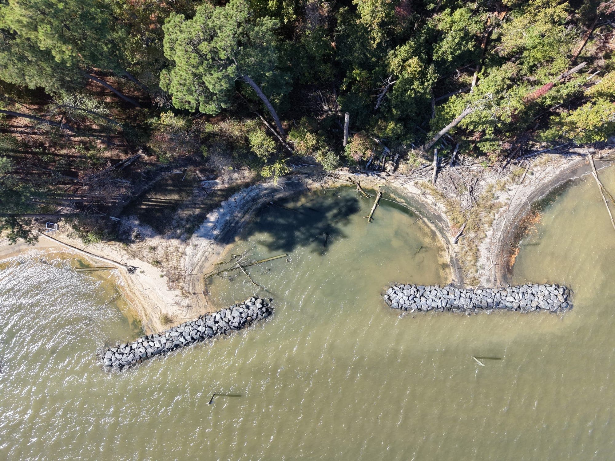 Top-down drone view of two stone breakwaters sheltering a pocket beach of sand fill below a forested bank at the Smithsonian Environmental Research Center.
