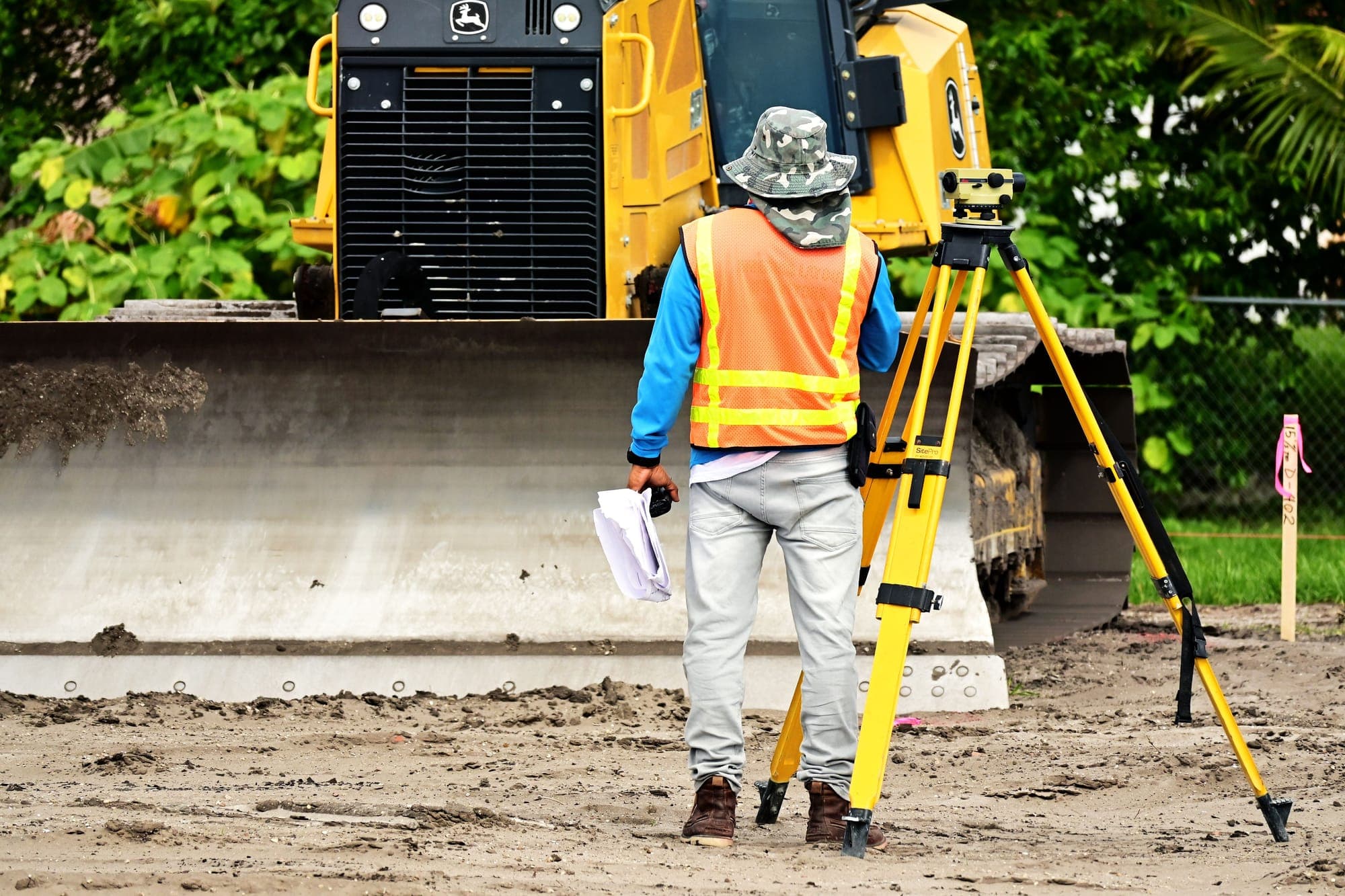 Surveyor in a safety vest holding plans beside a survey instrument on a tripod, with a bulldozer grading the site behind.