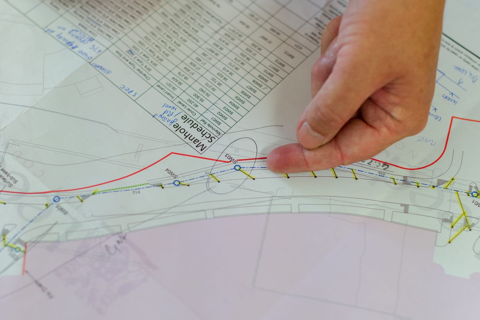 A hand pointing to a pipe alignment and manhole schedule on a civil site plan sheet.
